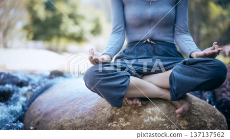 Person meditating in lotus pose on a boulder by a flowing river, calm outdoor nature scene with soft focus background. Person meditating in lotus pose on a boulder by a flowing river, calm outdoor nature scene with soft focus background. 137137562