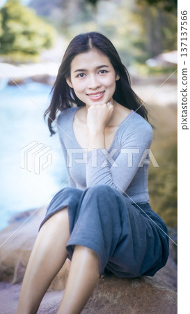 Smiling young woman sitting by a river outdoors, natural light, shallow depth of field, relaxed lifestyle portrait with copy space. Smiling young woman sitting by a river outdoors, natural light, shallow depth of field, relaxed lifestyle portrait with copy space. 137137566