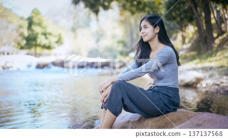 Young woman sitting on a riverside rock, smiling and relaxing in nature with soft morning light and shallow depth of field, outdoor lifestyle and mindfulness concept Young woman sitting on a riverside rock, smiling and relaxing in nature with soft morning light and shallow depth of field, outdoor lifestyle and mindfulness concept 137137568