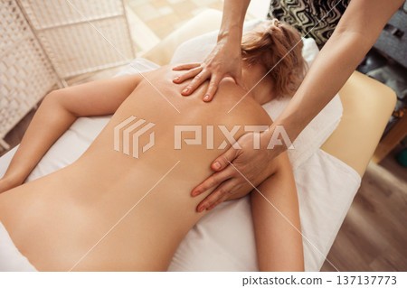 Overhead view of a spa therapist giving a soothing back massage to a young blonde woman on a white towel, warm soft light and neutral tones creating a calm wellness mood. 137137773