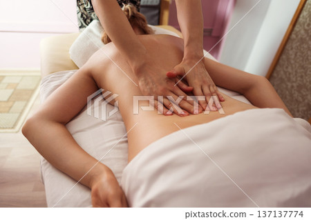 Close-up of a spa therapist hands gliding over a young woman bare back during relaxing massage, warm soft light highlighting smooth skin, cozy treatment room and tranquil wellness atmosphere. 137137774
