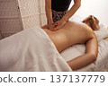 Spa therapist massaging the lower back of a young woman lying under a white sheet, warm natural light on smooth skin, wicker partition and soft textiles creating tranquil wellness atmosphere. 137137779