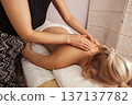 Relaxing spa massage with young blonde woman lying face down on white towel, receiving soothing back treatment from a therapist in patterned pants under warm, soft indoor light 137137782