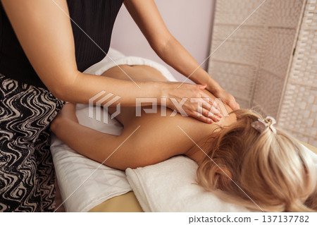 Relaxing spa massage with young blonde woman lying face down on white towel, receiving soothing back treatment from a therapist in patterned pants under warm, soft indoor light Relaxing spa massage with young blonde woman lying face down on white towel, receiving soothing back treatment from a therapist in patterned pants under warm, soft indoor light 137137782