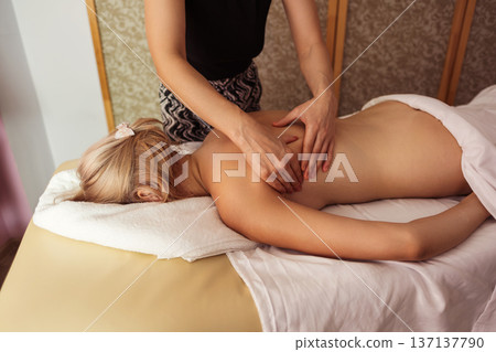 Close-up view of professional massage therapist performing back massage on a blonde woman lying on table, in spa room with warm light, calm tones, and relaxing atmosphere. 137137790