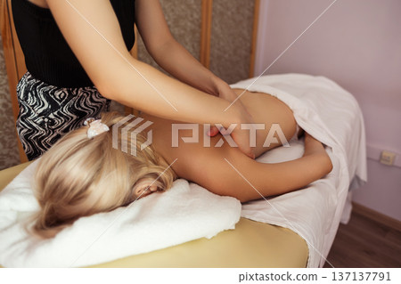Close-up view of professional massage therapist performing back massage on a blonde woman lying on table, in spa room with warm light, calm tones, and relaxing atmosphere. 137137791