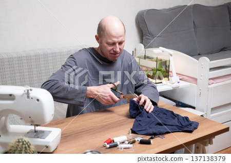 A bald man in his forties wearing a grey T-shirt sits at a table and cuts fabric with sewing scissors. A bald man in his forties wearing a grey T-shirt sits at a table and cuts fabric with sewing scissors. 137138379