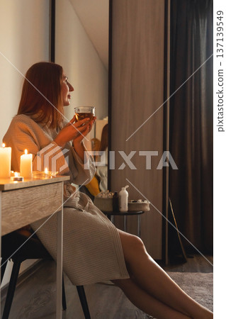 Woman in bathrobe enjoying hot tea illuminated by flickering candles, finding peace and tranquility. Wellness and spa evening Woman in bathrobe enjoying hot tea illuminated by flickering candles, finding peace and tranquility. Wellness and spa evening 137139549