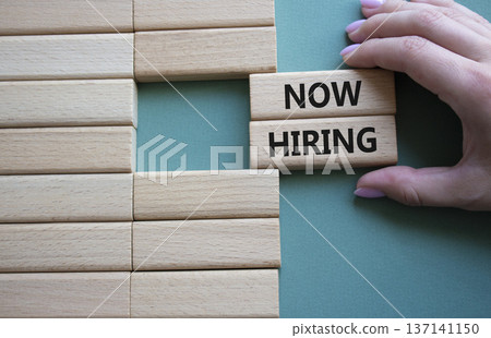 Now Hiring symbol. Concept word Now Hiring on wooden blocks. Businessman hand. Beautiful grey green background. Business and Now Hiring concept. Copy space 137141150