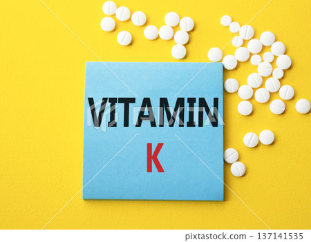 Vitamin K symbol. Concept word Vitamin K on blue note. Beautiful yellow background with pills. Medicine and Vitamin K concept. Copy space Vitamin K symbol. Concept word Vitamin K on blue note. Beautiful yellow background with pills. Medicine and Vitamin K concept. Copy space 137141535