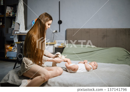 Mother gently puts on pants for her child lying on the bed Mother gently puts on pants for her child lying on the bed 137141796