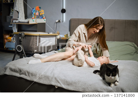 Mother plays with baby on the bed while cat walks around in a bedroom during the day Mother plays with baby on the bed while cat walks around in a bedroom during the day 137141800