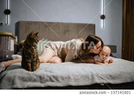 Mother and baby spend time together on bed with cat nearby in cozy bedroom setting Mother and baby spend time together on bed with cat nearby in cozy bedroom setting 137141801