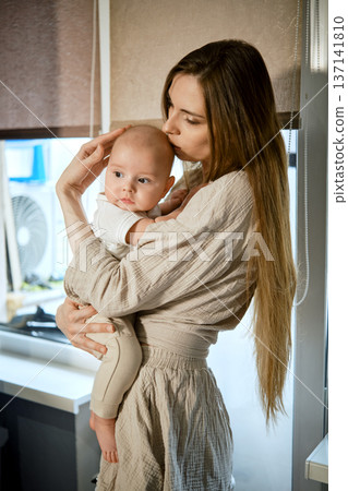 Mother holds baby while standing in home with soft lighting during daytime Mother holds baby while standing in home with soft lighting during daytime 137141810