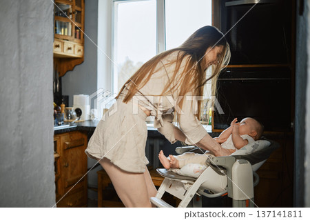 Mother interacts with baby in kitchen while preparing food during daytime Mother interacts with baby in kitchen while preparing food during daytime 137141811