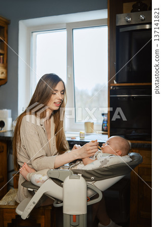 Mother feeds baby in a cozy kitchen during daytime while sitting together 137141821