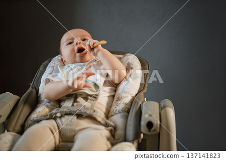 Baby eats with a spoon in a high chair during mealtime at home in the afternoon 137141823