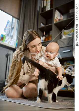 Mother and baby interact with a cat in a cozy living room during the day Mother and baby interact with a cat in a cozy living room during the day 137141836