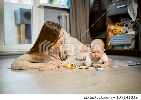 Mother plays with baby on the floor in a well-lit living room filled with toys and soft furnishings Mother plays with baby on the floor in a well-lit living room filled with toys and soft furnishings 137141839