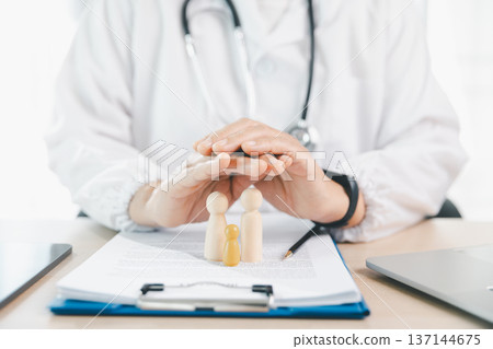 Doctor in white coat shields wooden family figures over clipboard paperwork with stethoscope and devices, symbolizing health insurance protection, patient safety and coverage planning. protection Doctor in white coat shields wooden family figures over clipboard paperwork with stethoscope and devices, symbolizing health insurance protection, patient safety and coverage planning. protection 137144675