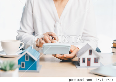 Mortgage and real estate budgeting concept with consultant using calculator beside miniature homes, coffee cup and paperwork on bright desk, symbolizing home loan planning and insurance. Mortgage and real estate budgeting concept with consultant using calculator beside miniature homes, coffee cup and paperwork on bright desk, symbolizing home loan planning and insurance. 137144683