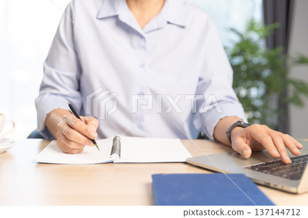 Business professional working on laptop at modern office desk with documents, smartphone and stationery, bright daylight by window. Concept of productivity, remote work and digital management. 137144712