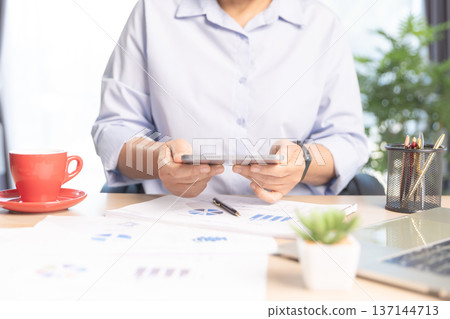 Business professional working on laptop at modern office desk with documents, smartphone and stationery, bright daylight by window. Concept of productivity, remote work and digital management. Business professional working on laptop at modern office desk with documents, smartphone and stationery, bright daylight by window. Concept of productivity, remote work and digital management. 137144713