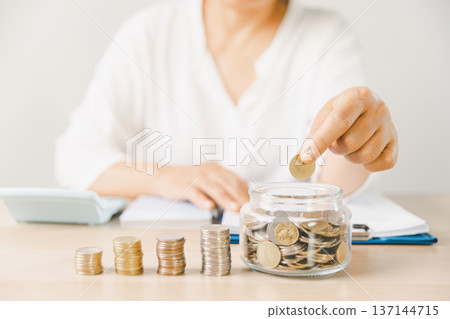 Close-up of hand putting coin into clear glass jar filled with change on bright desk. Minimal home scene for personal savings, budgeting, money management, future goals and security. money management Close-up of hand putting coin into clear glass jar filled with change on bright desk. Minimal home scene for personal savings, budgeting, money management, future goals and security. money management 137144715