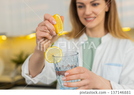 Middle-aged woman squeezing lemon into water glass in home kitchen for detox hydration daily routine Middle-aged woman squeezing lemon into water glass in home kitchen for detox hydration daily routine 137146881