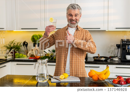 Middle-aged man drinking lemon water giving thumbs up in kitchen for healthy fitness diet hydration Middle-aged man drinking lemon water giving thumbs up in kitchen for healthy fitness diet hydration 137146928
