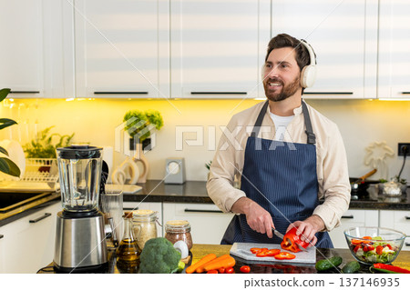 Middle-aged man singing with headphones while slicing red pepper for healthy salad in home kitchen 137146935