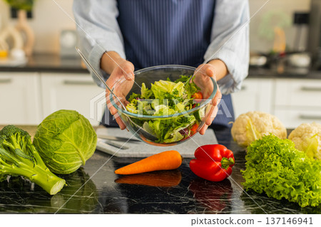 Woman holding glass bowl of fresh vegetable salad on kitchen counter for healthy lunch meal concept Woman holding glass bowl of fresh vegetable salad on kitchen counter for healthy lunch meal concept 137146941