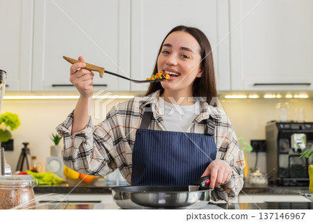 Young woman tasting cooked vegetables from pan in kitchen testing flavor for healthy home dinner hot Young woman tasting cooked vegetables from pan in kitchen testing flavor for healthy home dinner hot 137146967