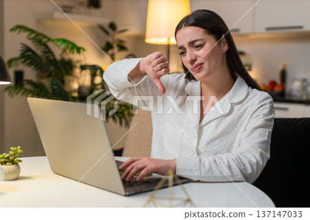 Young woman loses online game on laptop drops shoulders shows thumbs down then tries again evening Young woman loses online game on laptop drops shoulders shows thumbs down then tries again evening 137147033
