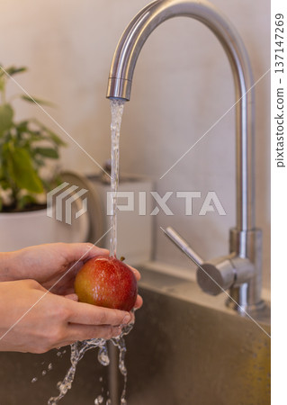 Young woman in kitchen with apron washes red apple under running tap water at sink clean fresh prep 137147269