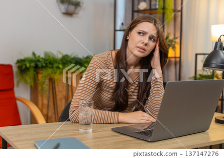 Young woman rubs temples after long laptop work drinks water squints from fatigue and breathes slow 137147276