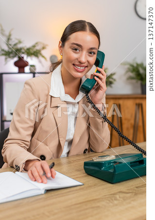 Young businesswoman answers retro landline from nineties speaks clear business talk and writes notes Young businesswoman answers retro landline from nineties speaks clear business talk and writes notes 137147370