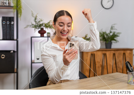 Young businesswoman typing fast on smartphone reads news of app game win and raises fist in victory Young businesswoman typing fast on smartphone reads news of app game win and raises fist in victory 137147376