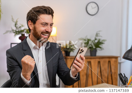 Middle-aged businessman texting happy news on smartphone celebrates project win with victory gesture Middle-aged businessman texting happy news on smartphone celebrates project win with victory gesture 137147389