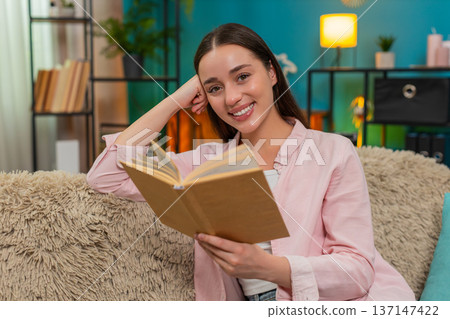 Young woman reading book on sofa turning pages slowly reflecting deeply on story absorbing meaning 137147422