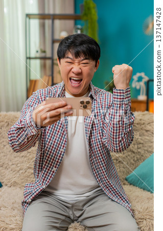 Asian man excitedly playing mobile game on smartphone, smiling loudly celebrating level victory Asian man excitedly playing mobile game on smartphone, smiling loudly celebrating level victory 137147428