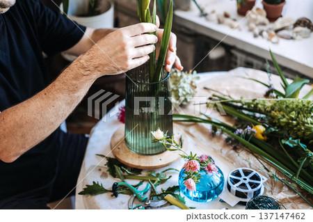 Hands of florist place long green stems into glass vase on table with tools and floral supplies. Independent florist, local business owner, small business startup, craft industry, flower shop studio. 137147462