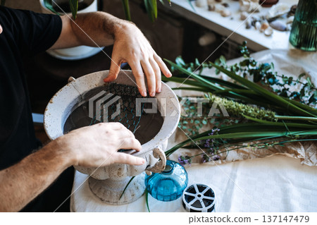 Hands place wire mesh into stone urn for floral arrangement preparation. Sustainable floral design, plastic-free floristry, eco-friendly flower arranging. Hands place wire mesh into stone urn for floral arrangement preparation. Sustainable floral design, plastic-free floristry, eco-friendly flower arranging. 137147479