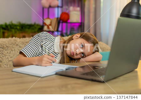 Child girl kid doing lazy homework at home, sitting tired at table with laptop during online school 137147844