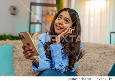 Indian woman feeling nostalgic while looking at old photos on smartphone at home, sits on soft sofa Indian woman feeling nostalgic while looking at old photos on smartphone at home, sits on soft sofa 137147916