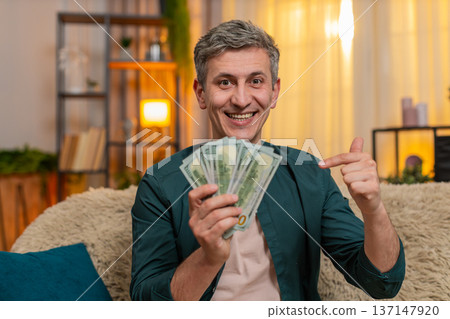 Young man smiles while counting cash dollars in hands expresses joy from income and financial gain Young man smiles while counting cash dollars in hands expresses joy from income and financial gain 137147920