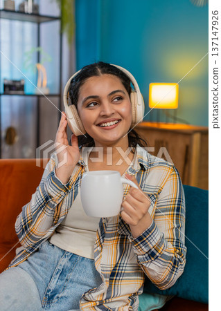 Indian woman wearing headphones listening calming relaxing music holding coffee cup carefree at home Indian woman wearing headphones listening calming relaxing music holding coffee cup carefree at home 137147926