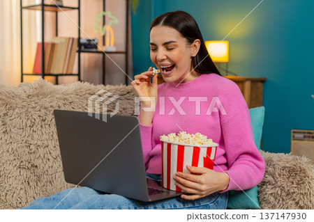 Young woman on sofa watching movie on laptop eating popcorn laughing enjoying film entertainment 137147930