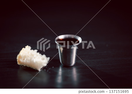Minimal still life of communion cup with red wine and bread on dark table, symbolizing Holy Communion, Eucharist, Christian faith, worship, sacrifice and remembrance with ample copy space. 137151695