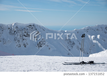 Skis and poles standing on snowy mountain ridge in Krasnaya Polyana with alpine peaks and blue sky in background. Minimal winter landscape suitable for tourism marketing, sport equipment Skis and poles standing on snowy mountain ridge in Krasnaya Polyana with alpine peaks and blue sky in background. Minimal winter landscape suitable for tourism marketing, sport equipment 137151852